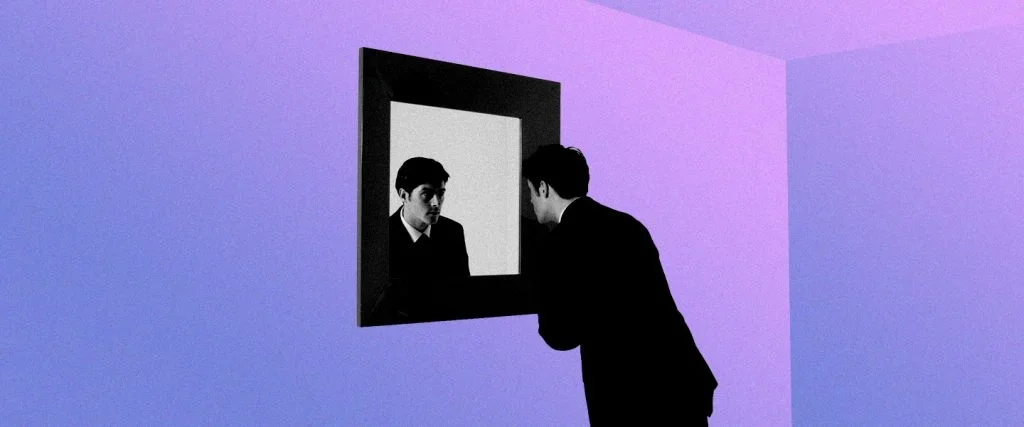 A man in a suit looking at his reflection in a mirror on a purple wall.