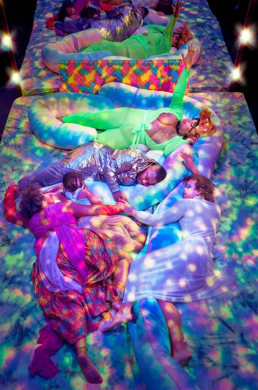 People lying on colorful beds in a psychedelic, rainbow-lit setting, with reflections in a mirror showing various poses and expressions.