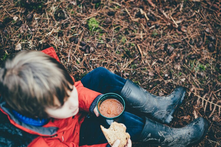 primary aged child sits on the ground in wellies with cup of hot chocolate