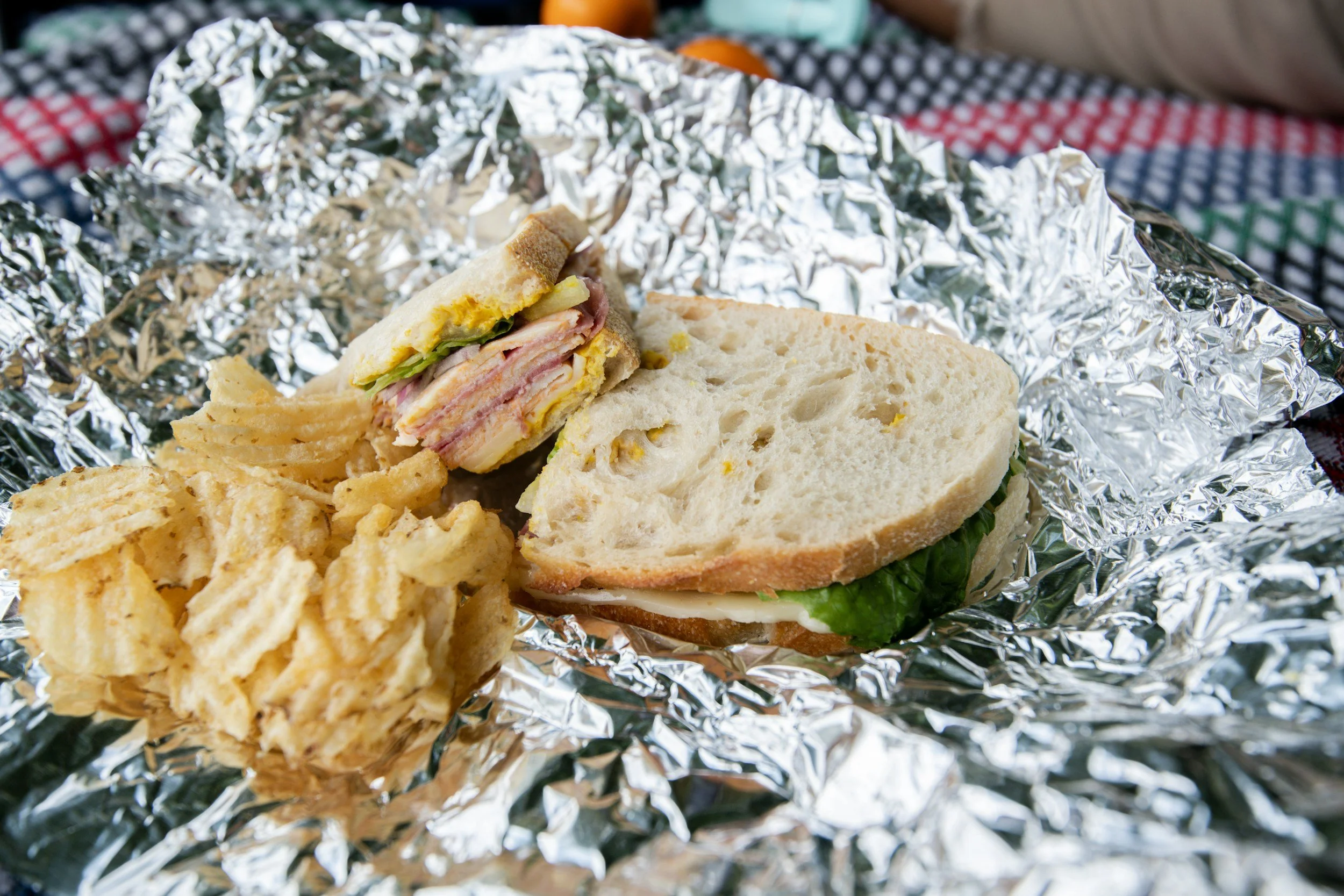 Image shows a white bread sandwich containing cold meats and salad with a pile of crisps, on an open foil wrapper.