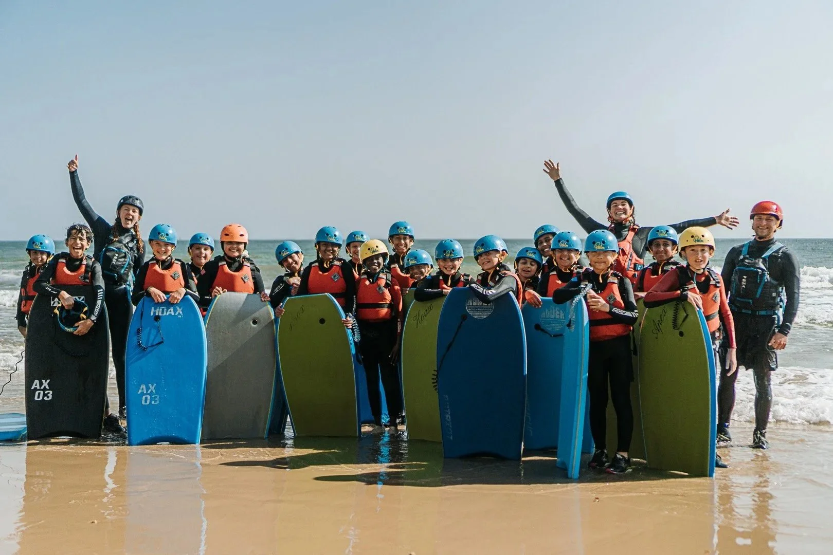 Image: Land & Wave School Trips. A group of children from Griffefield Primary School are standing on a beach with the sea behind them. They are wearing wetsuits and red buoyancy aids and holding blue body boards. Three instructors are with the group.