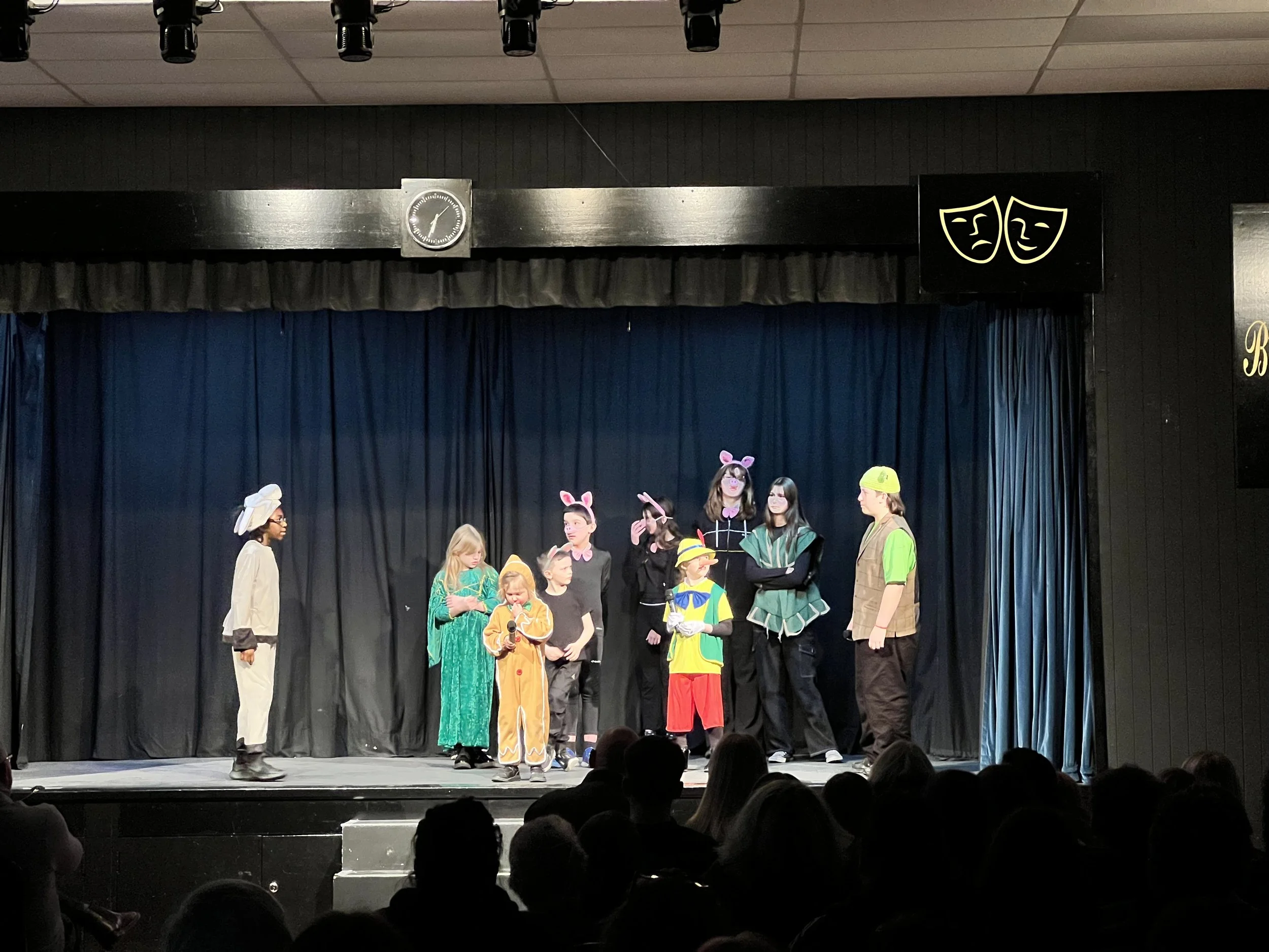 Children performing a play on stage dressed in costumes like animals, a farmer, and a fairy tale characters. The stage has a dark curtain backdrop and a clock above.