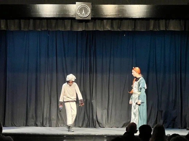 Two young girls dressed in costumes performing on a stage with a blue curtain backdrop. One girl is dressed as a chef, and the other as a queen or princess. There is a clock on the wall above the stage.