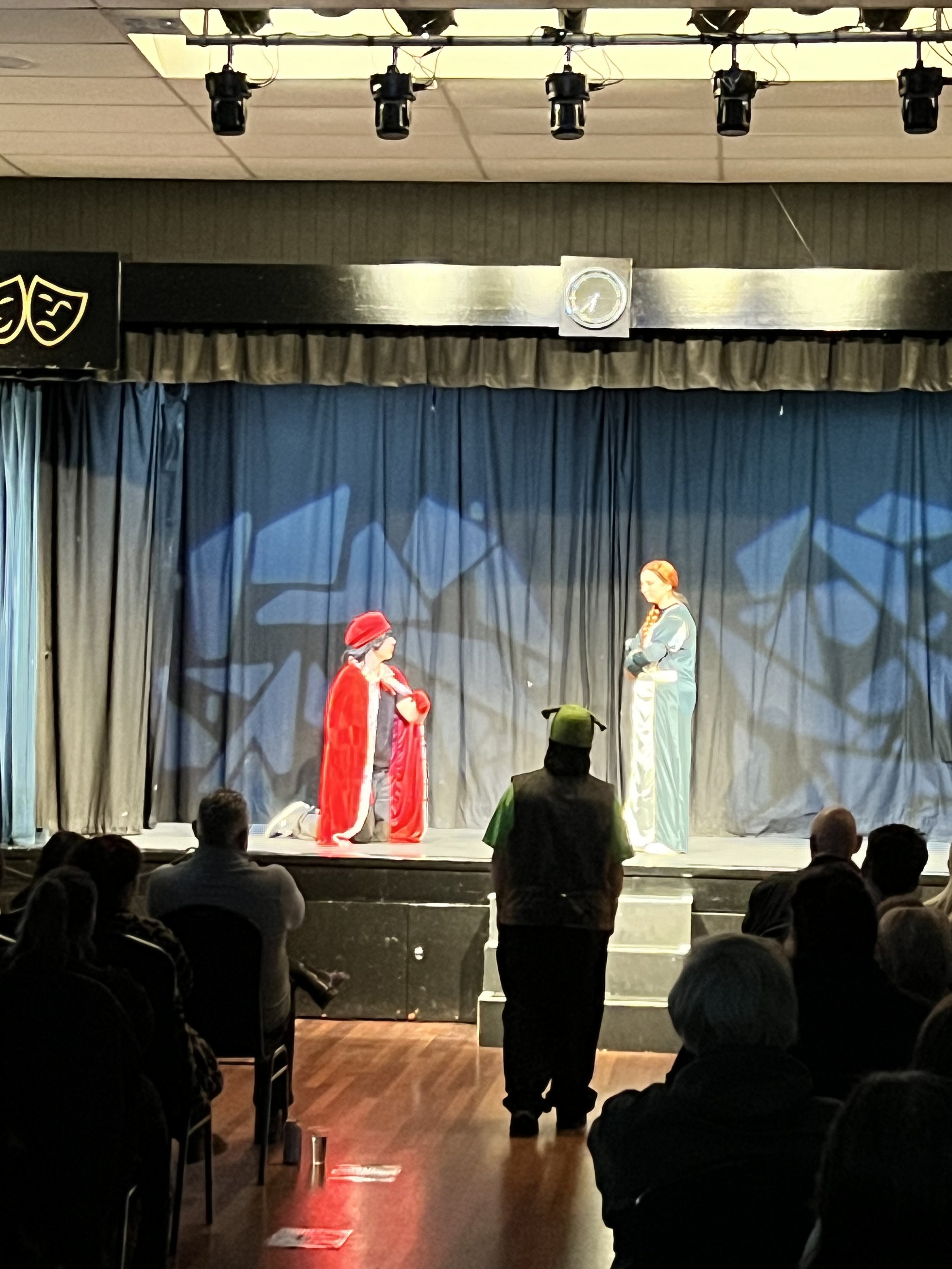 A stage with two actors performing a play, with one in a red cloak and hat kneeling, and the other in a blue patterned outfit, in front of an audience.