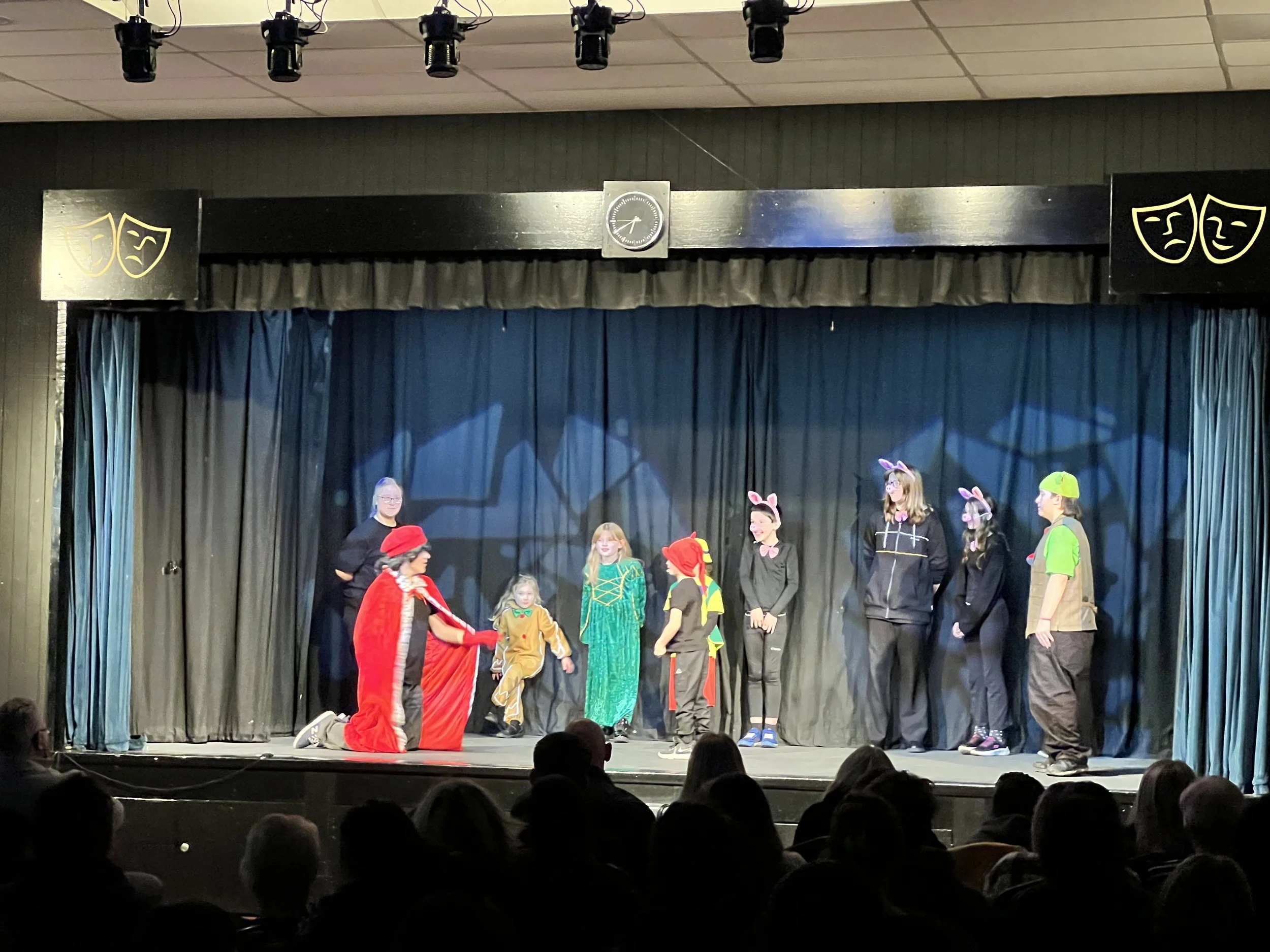 Children performing a play on stage, dressed in costumes including animals and a princess, with an audience watching.