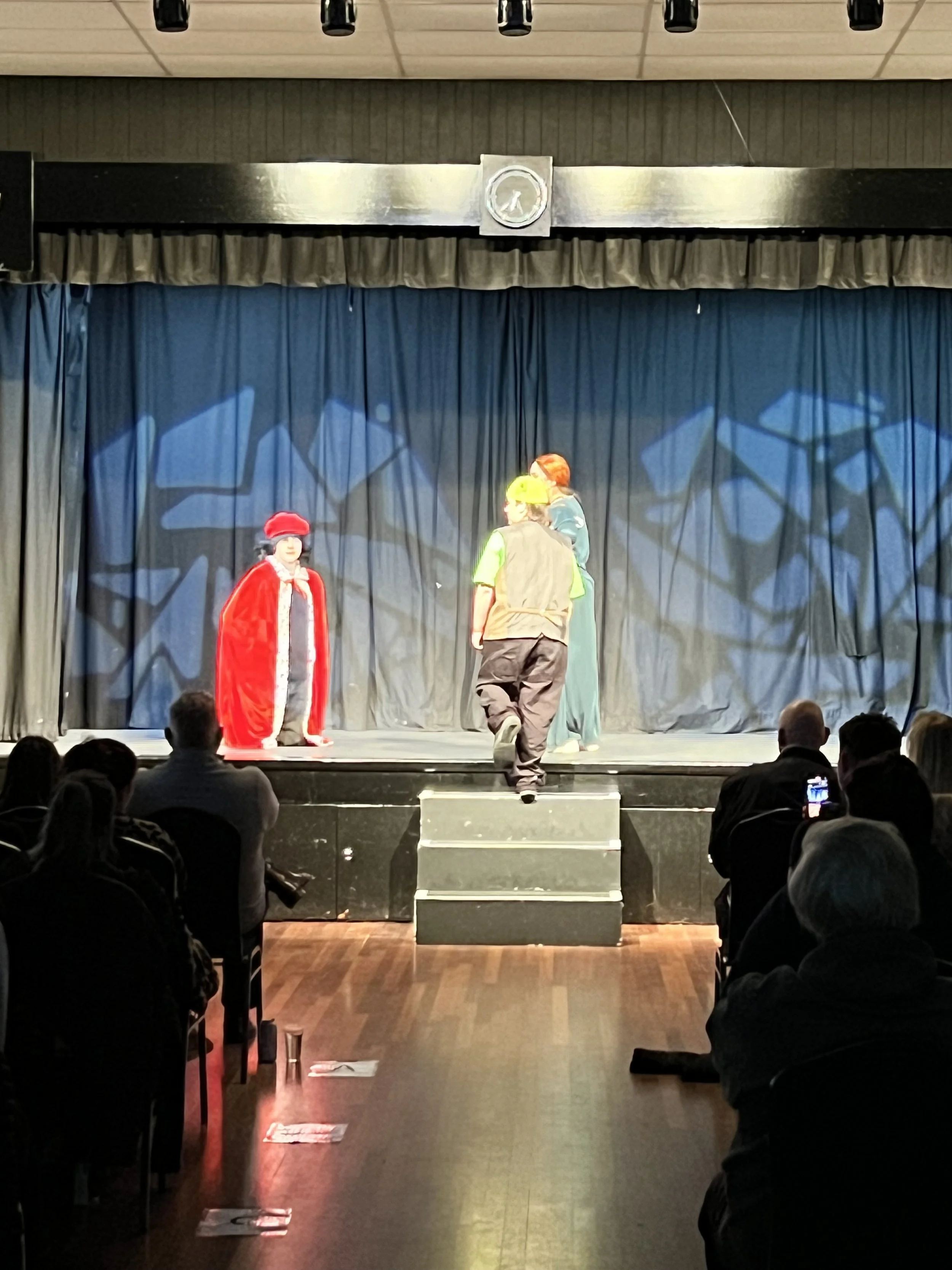 Children performing a play on stage with a black curtain backdrop, audience seated in front, stage lights overhead, and stage steps leading up to the performers.