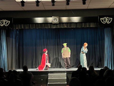 Three children performing a play on a stage with a dark curtain, on a school auditorium stage with audience silhouettes visible.
