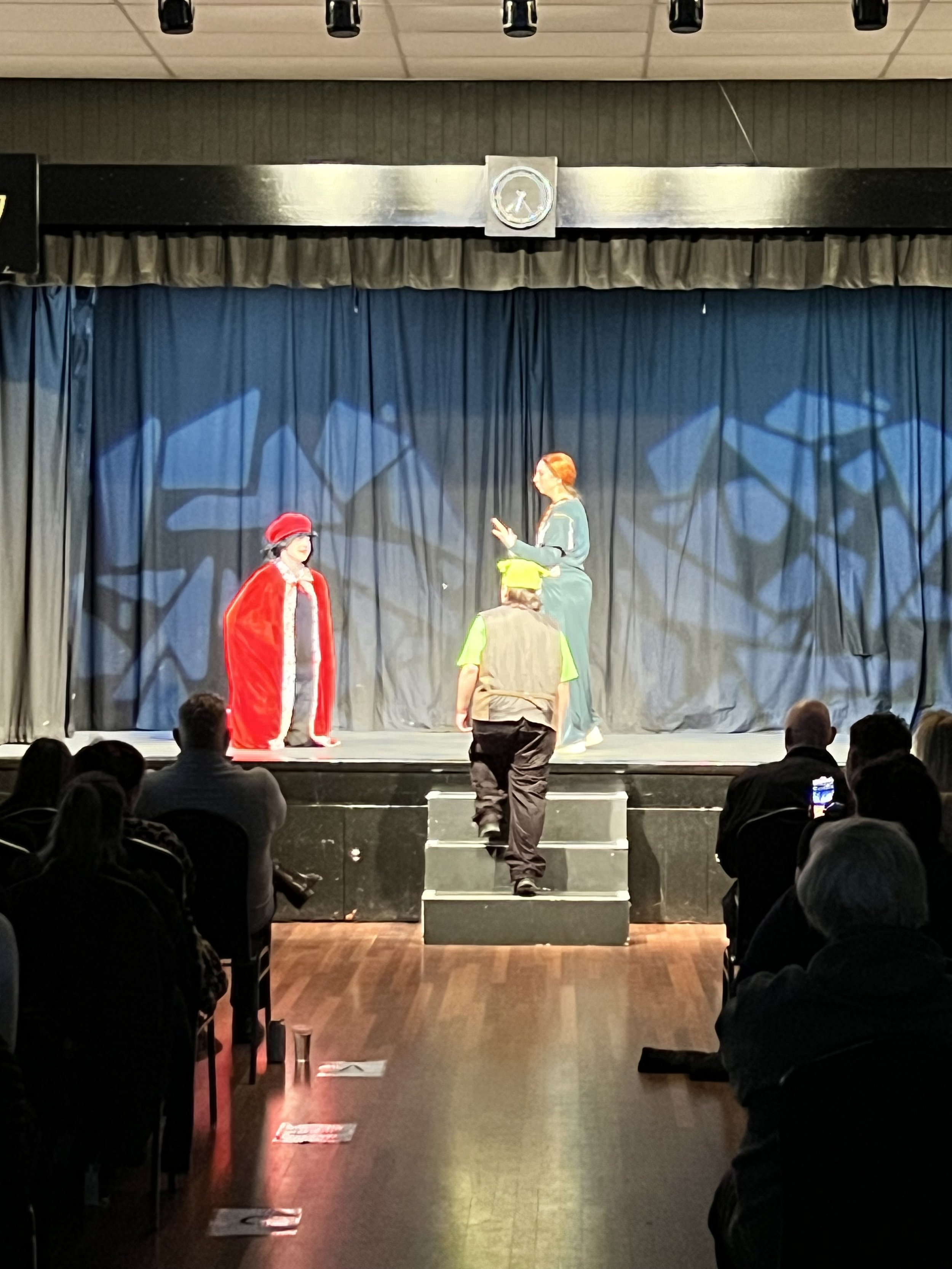 Children performing a play on stage with an adult, under stage lighting, with an audience watching.