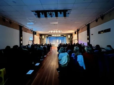 Audience seated in a dark auditorium watching a presentation or performance on stage with curtains and lighting.