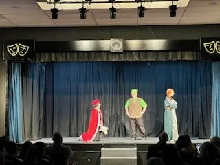 Three actors on stage performing in a theater with a blue curtain backdrop, audience in foreground