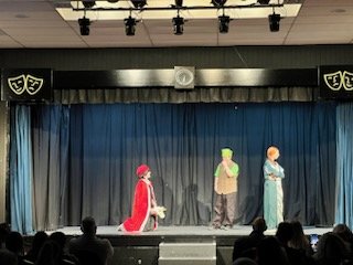 Three children in costumes perform on a stage with a blue curtain backdrop, audience visible in front.