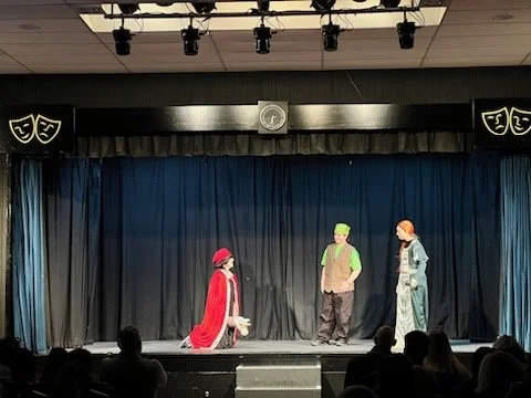 Theater stage with three actors in costume performing in front of an audience, black curtains, and theatrical shields on the side walls.