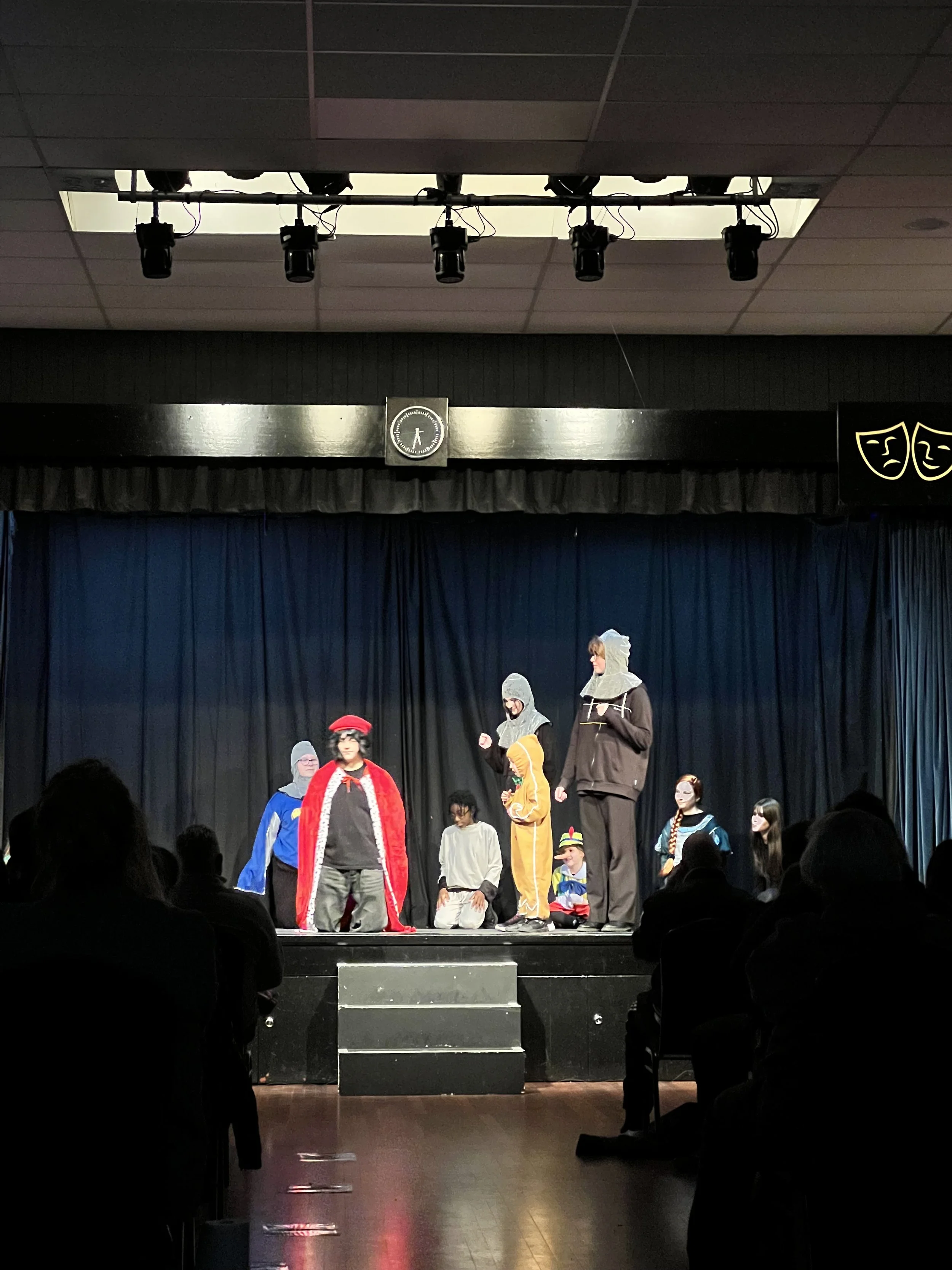 Children dressed in colorful costumes performing on stage with a blue curtain backdrop; audience seated in front.