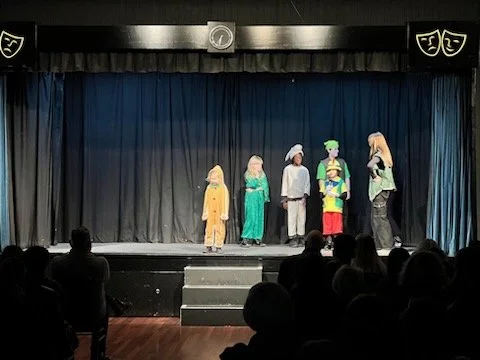 Children performing on a stage in a school play with blue curtains and theatrical masks above, audience seated in front.