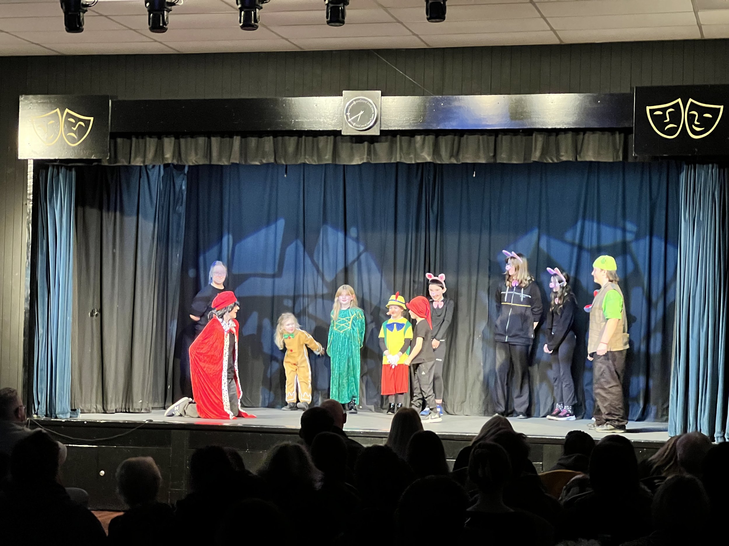 Children performing a play on stage dressed in colorful costumes, including animals and storybook characters, with an audience watching.