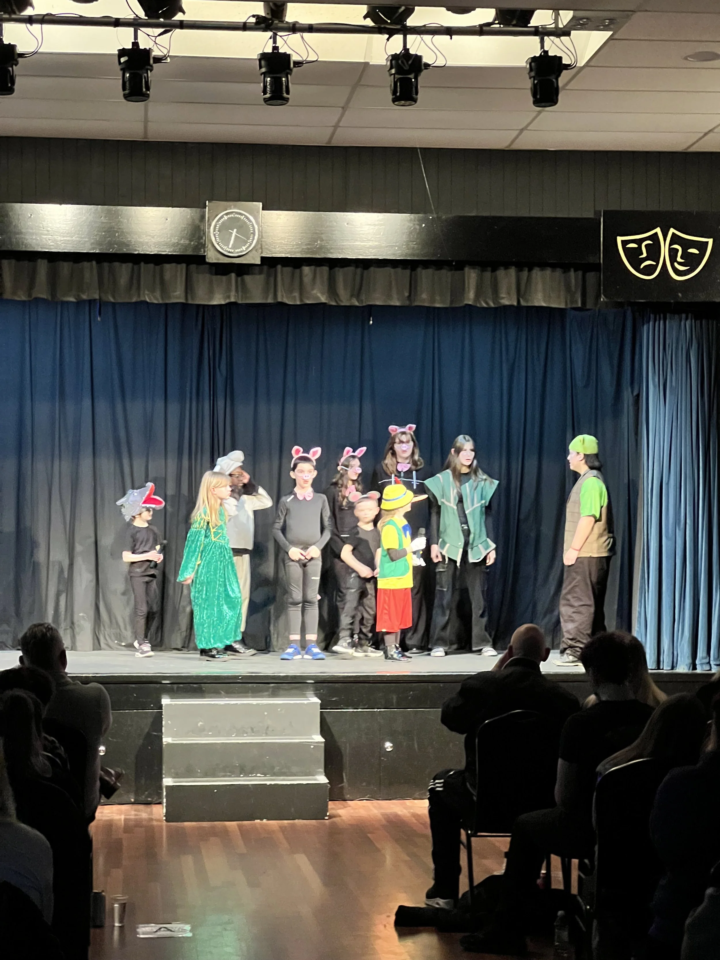 Children performing a play on stage with costumes, including animals and a fisherman, with an audience watching in a darkened theater.