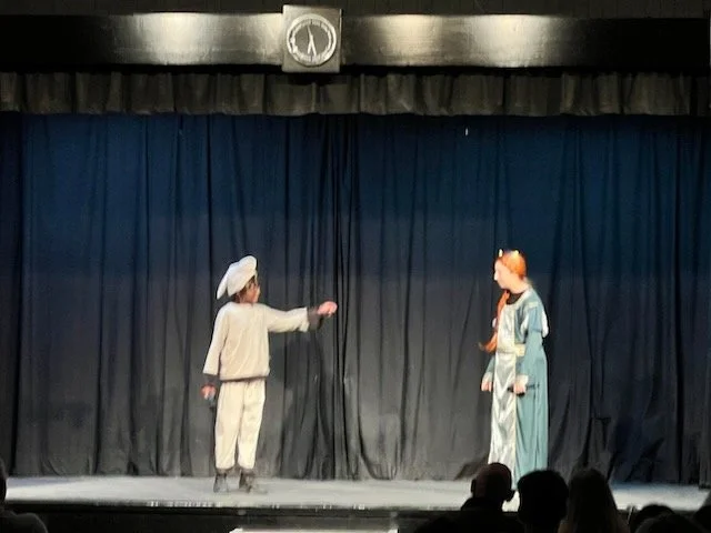 Two children performing on stage in costumes, one dressed as a nurse and the other as a soldier, in front of a black curtain with a clock above.