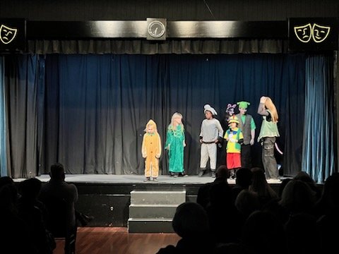 Children performing a play on a stage with a black curtain backdrop, dressed in colorful costumes including a lion, a princess, and other characters.