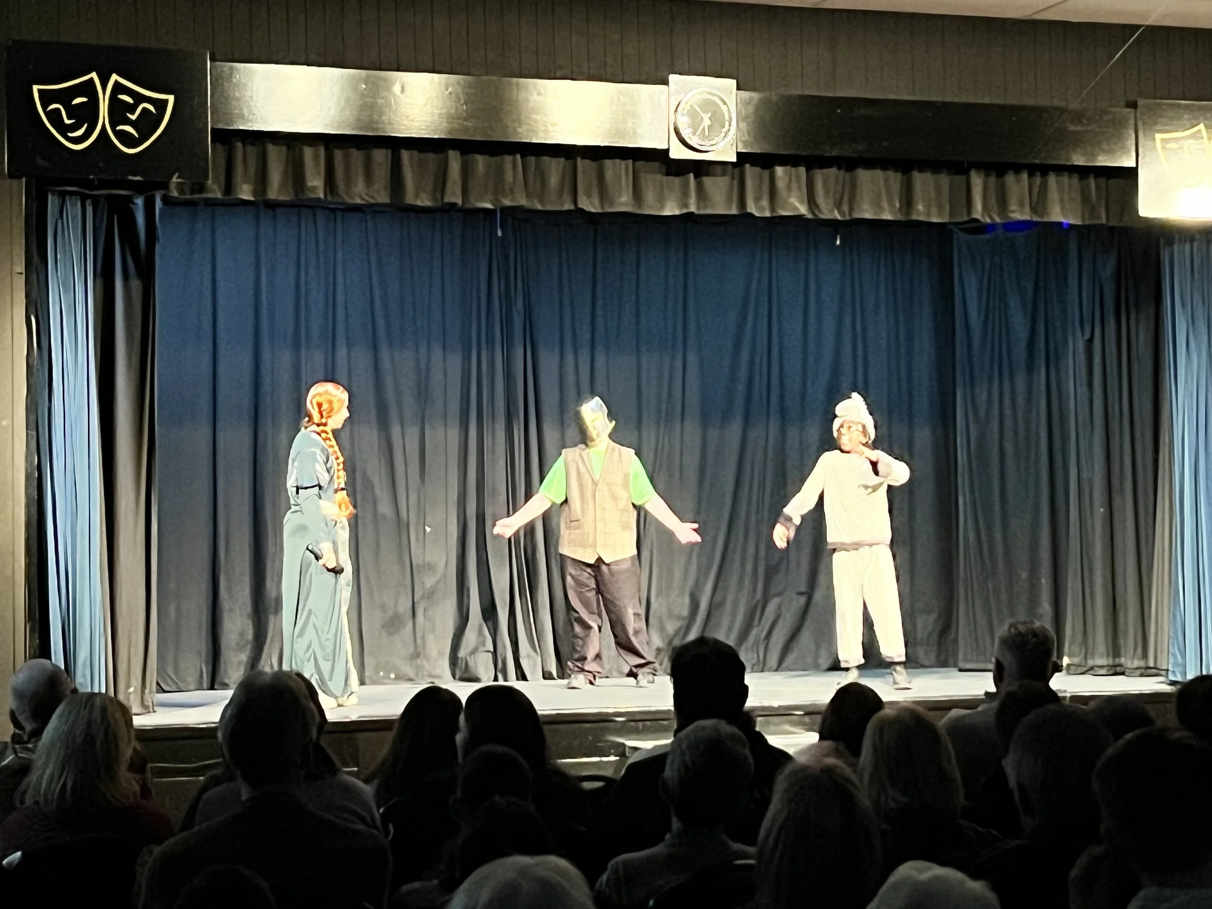 Three performers on stage in costume with a dark curtain background, audience watching in foreground.