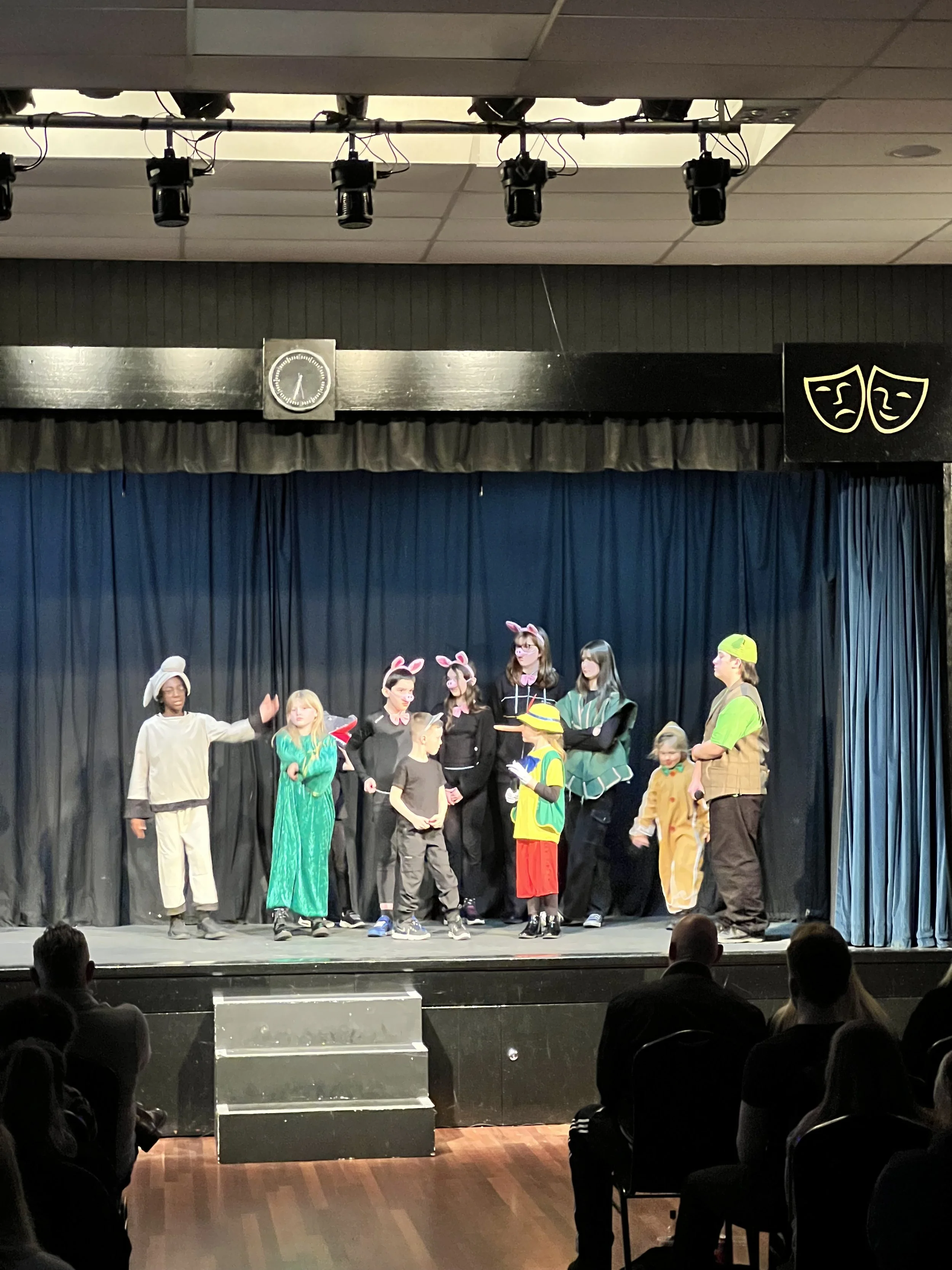 Children performing a play on stage, dressed in various costumes including animals, a bunny, and a person with a walking stick, with an audience watching.
