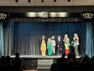 Children performing a play on a stage with a blue curtain, some in costumes and others in casual clothing.