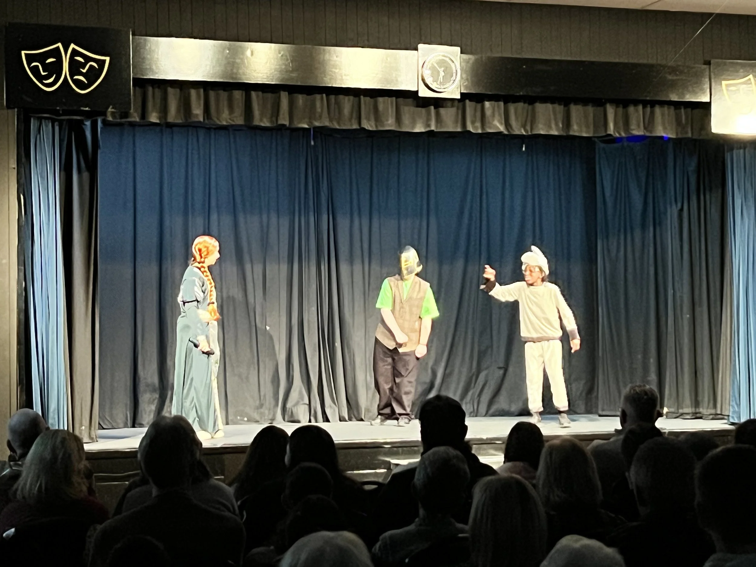 A theater stage with three performers dressed in costumes, engaging in a play, with an audience watching.