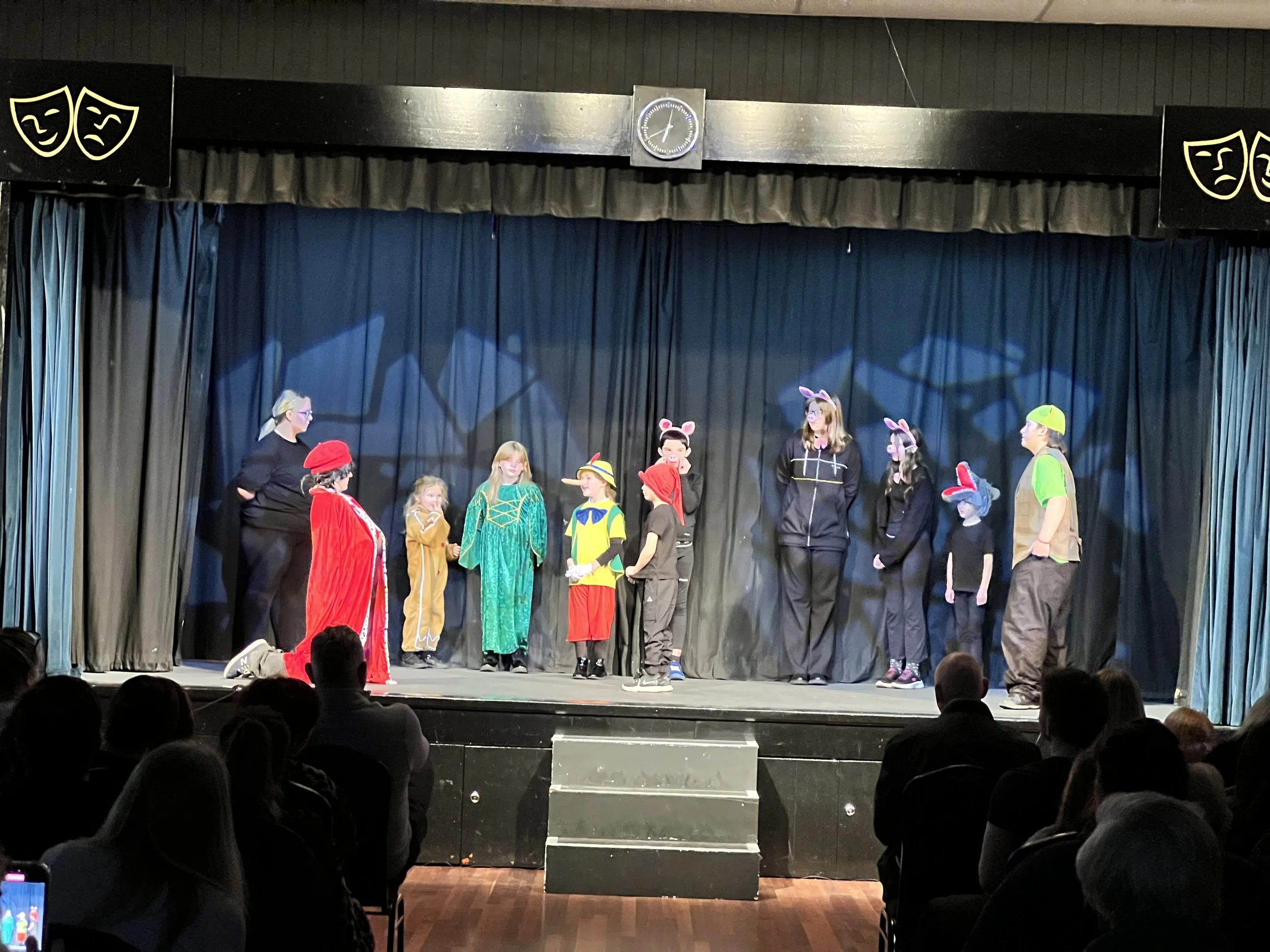 Children performing a play on stage dressed in various animal and character costumes, with an audience watching.