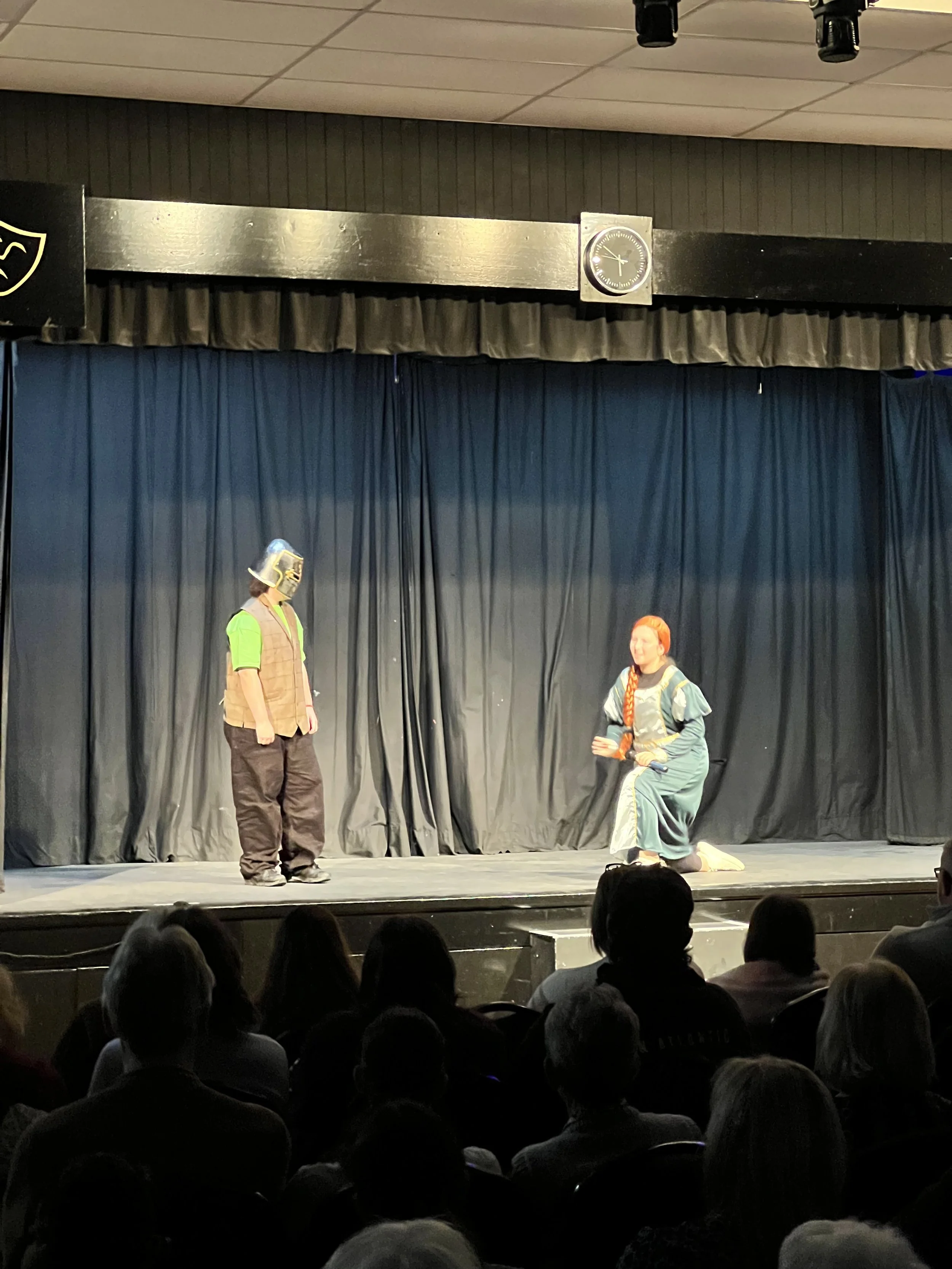 Two performers on a stage with a dark curtain in the background; one standing wearing a helmet and casual clothing, the other kneeling in a long dress, holding a prop, engaging in a theatrical performance, with an audience watching.