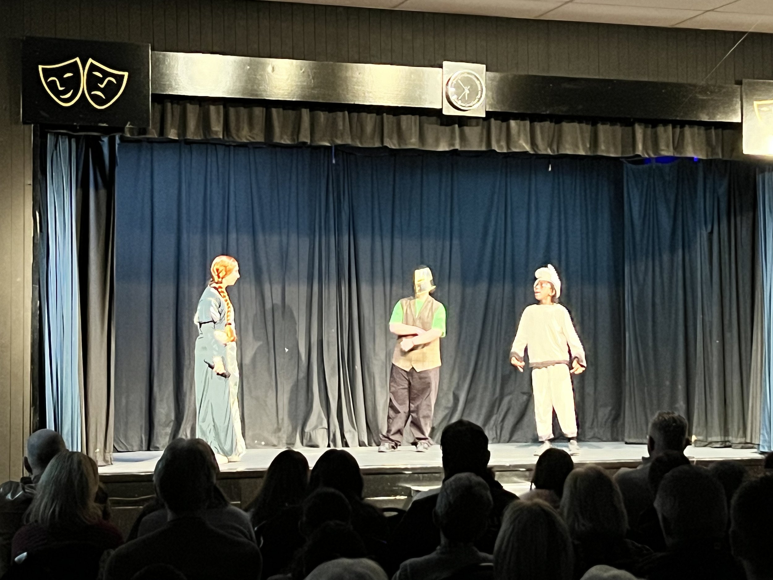 Children performing a play on a stage with black curtains, an audience watching, theater masks logo on the top left, and a clock above the stage.