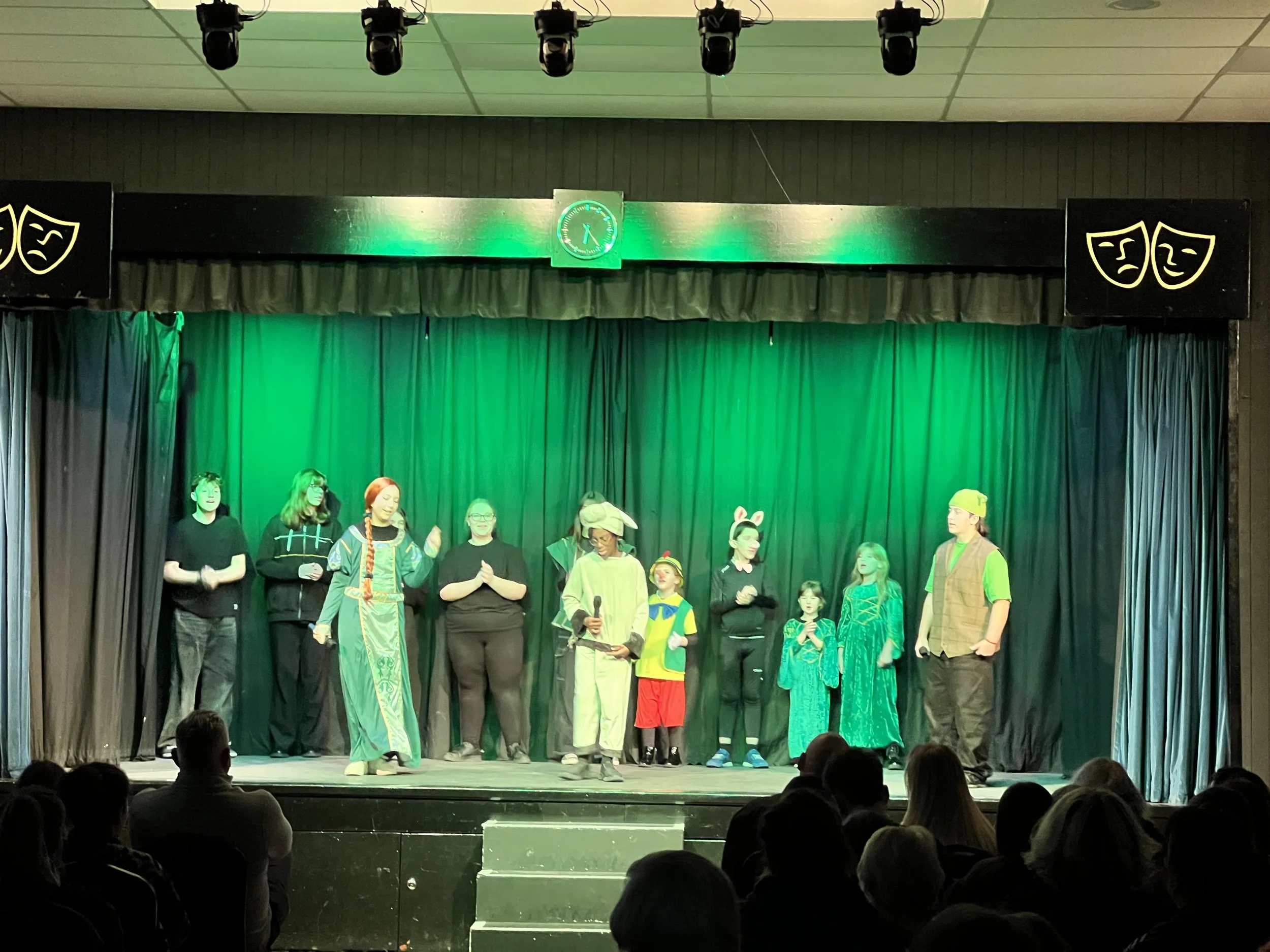 Children and adults perform a play on stage, with some dressed in costumes such as animals and fairy-tale characters, under green lighting, in front of an audience.