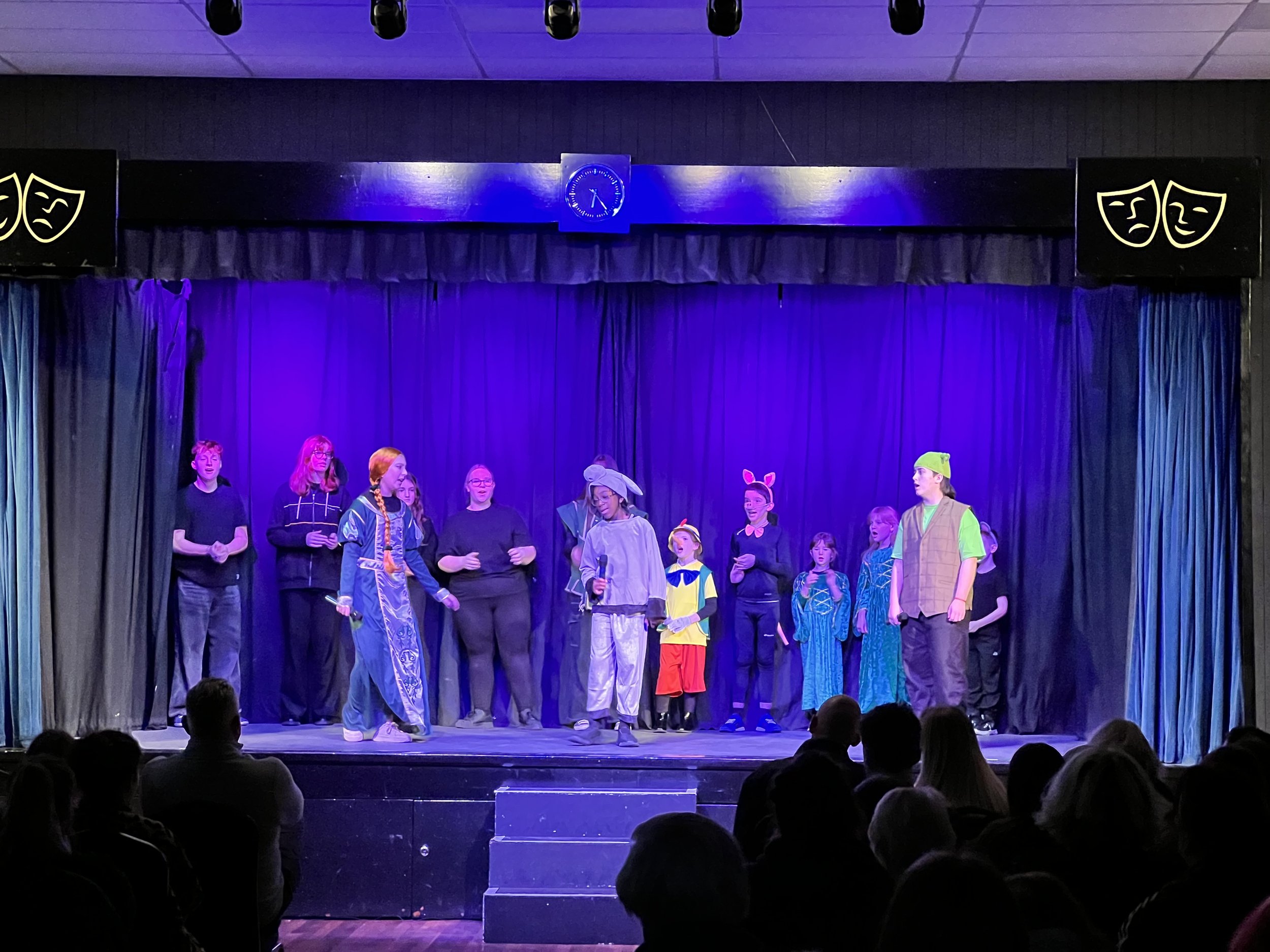 Children performing a play on stage with purple curtains and masks symbols, audience watching in a darkened theater.