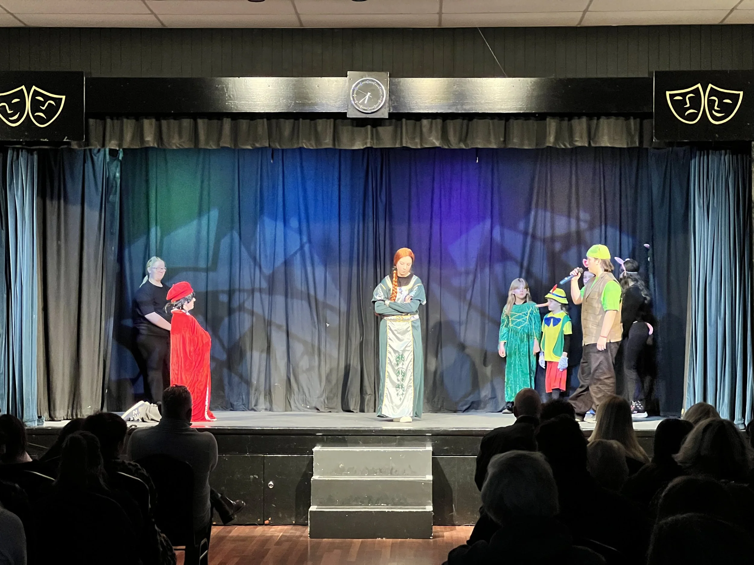 Children and an adult on stage performing a play, with audience watching. Stage has colorful costumes, darker curtains, theatre icons and a clock at the top.