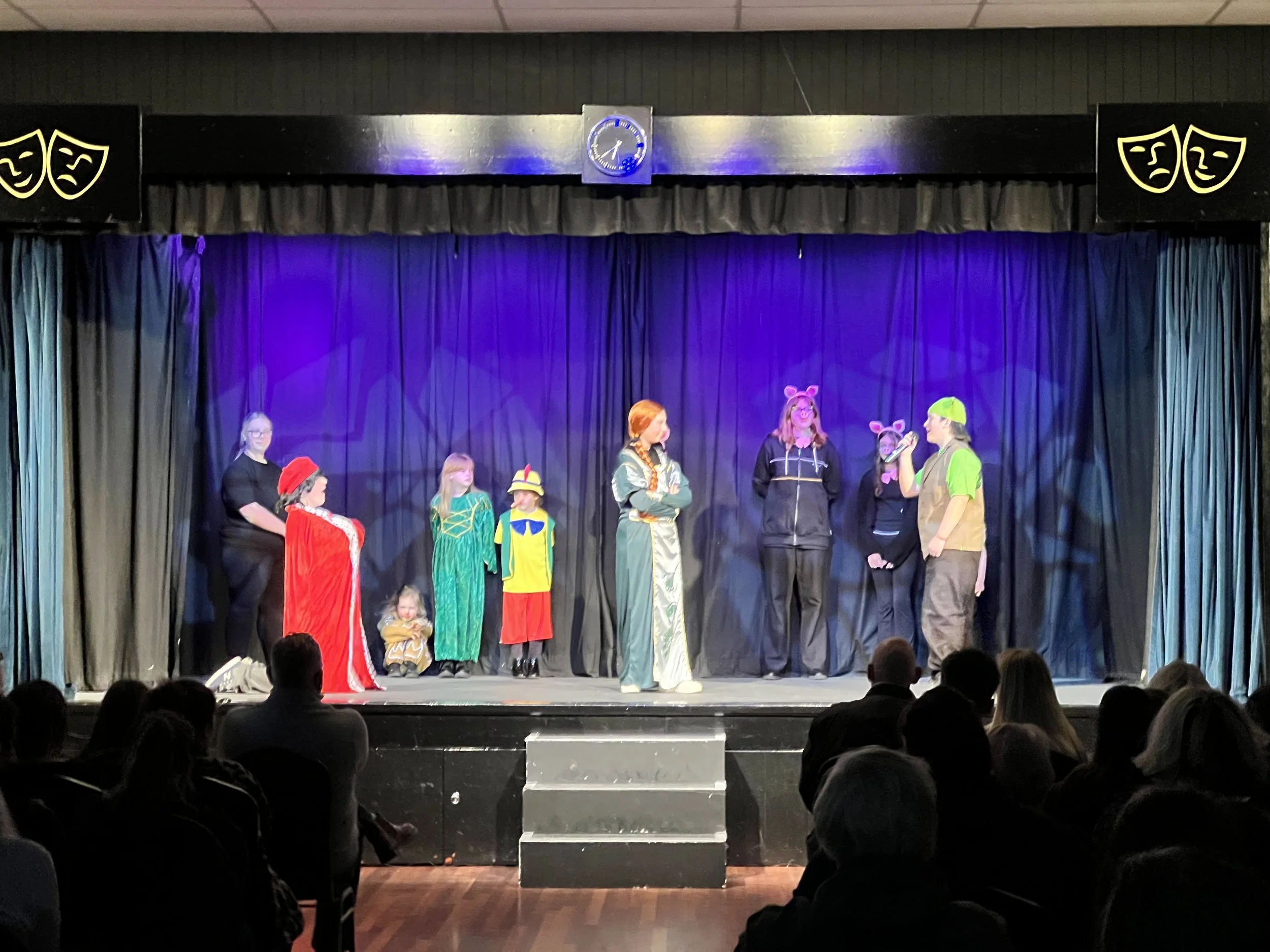 Children and adults on stage performing a play, with some dressed in costumes like animals and characters, with a black curtain background, stage lights, and an audience watching.