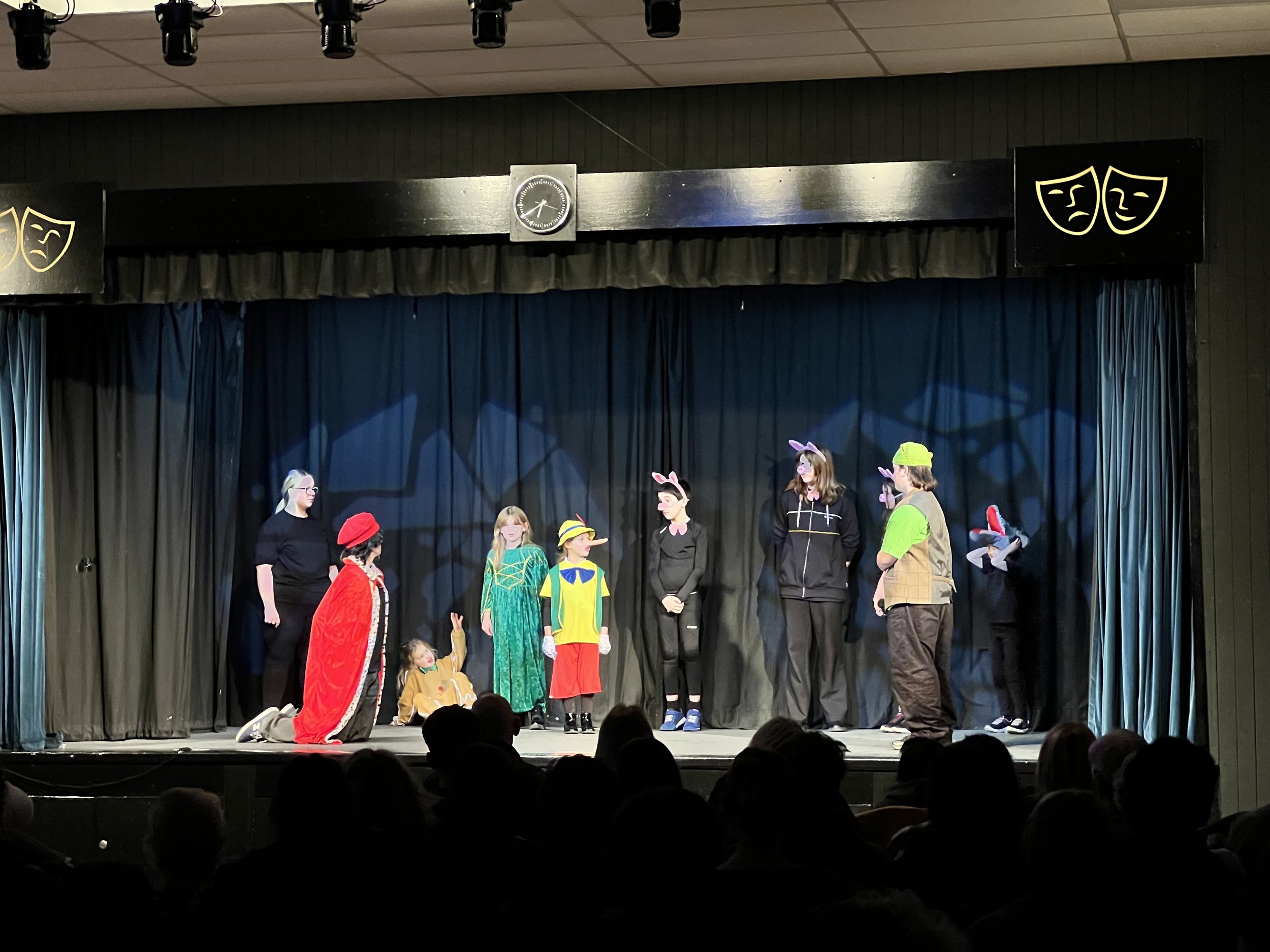 Children and adults performing a play on stage dressed in costumes including animals, a princess, and a baseball player, with a black curtain background and theatre lighting.