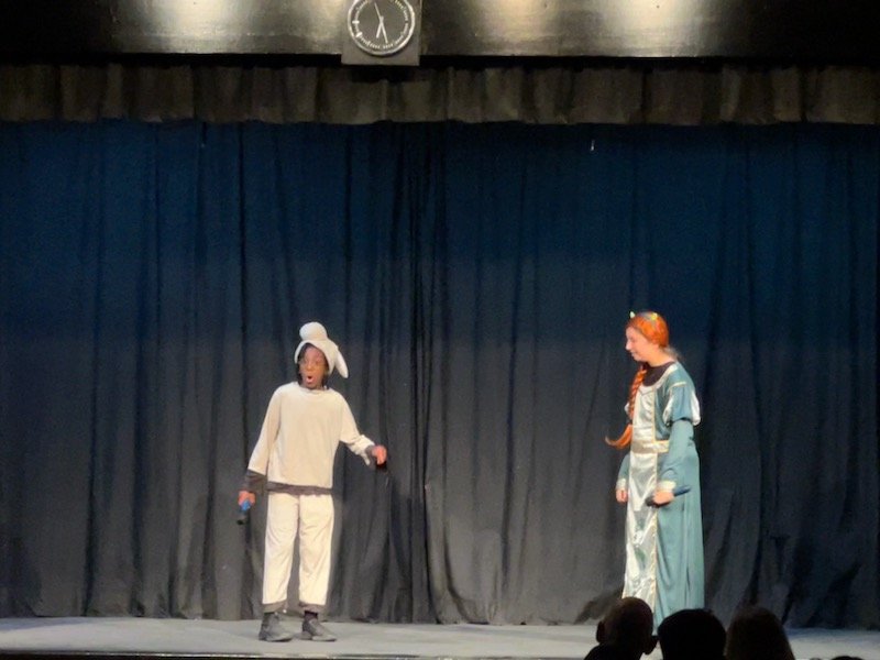 Two children performing on a stage with a black curtain backdrop, one dressed as a rabbit and the other as a princess, under a clock showing 12:20.