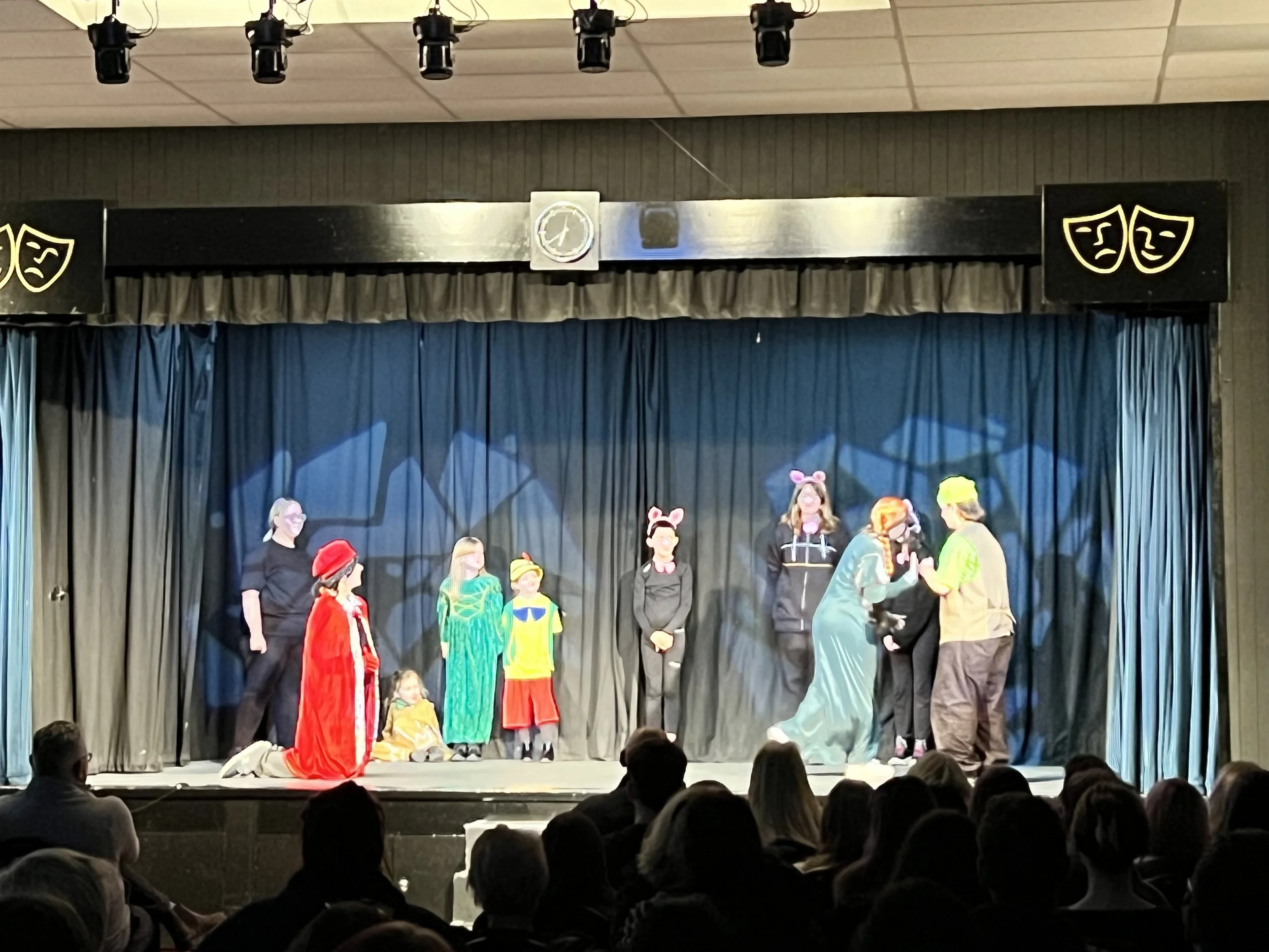 Children performing a play on stage with costumes, puppets, and an audience watching.