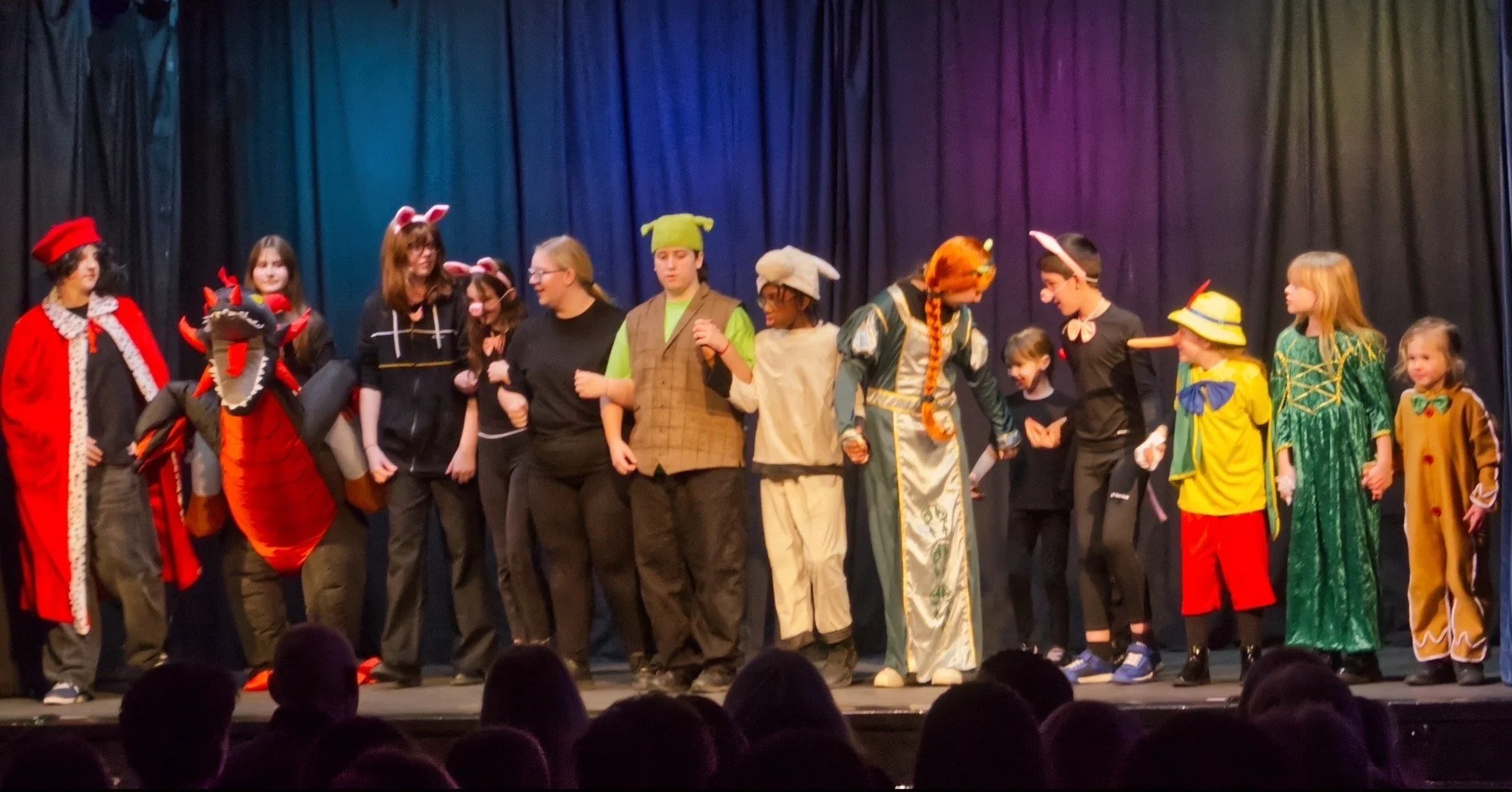 A group of children in costume taking a bow on stage after a performance, with a black curtain backdrop and the audience in the foreground.