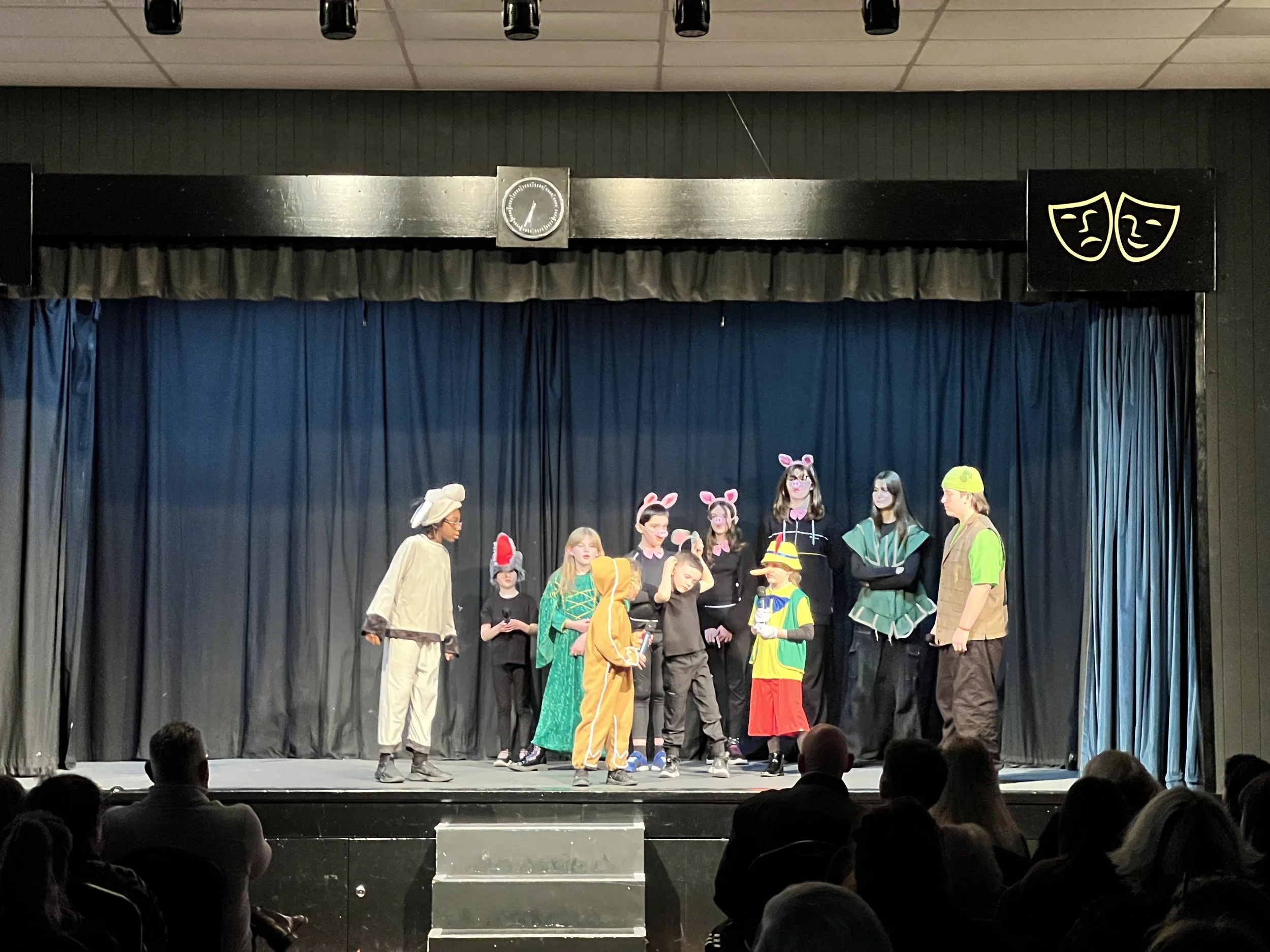 Children performing a play on stage dressed in various costumes including animals, a clown, and fairy tale characters, with an audience watching.