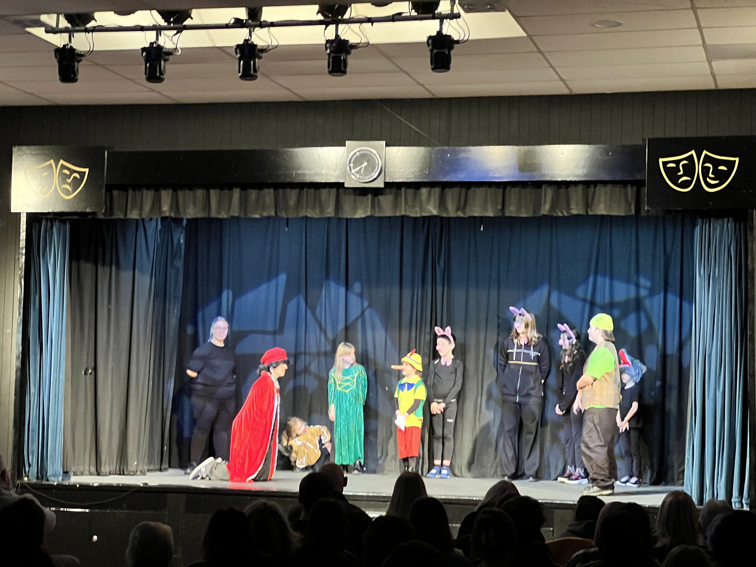 Children performing a play on stage with black curtains, some dressed as animals and characters, with audience members watching from below.