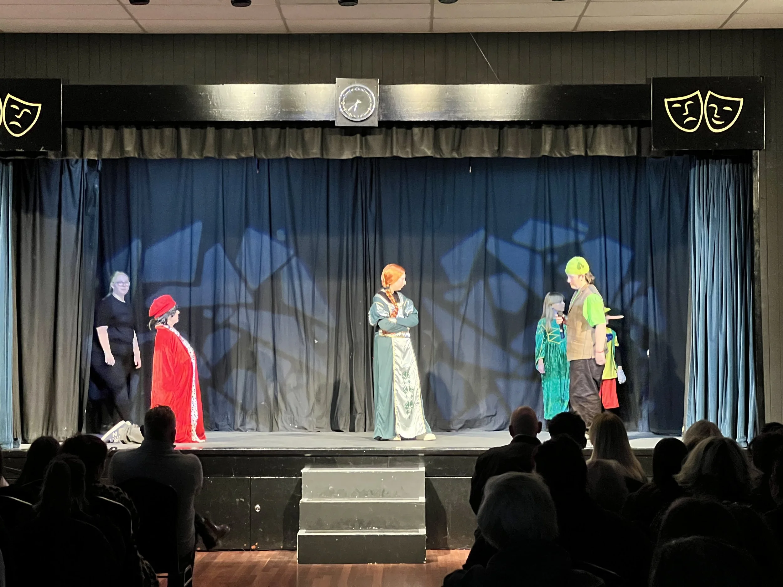 Children performing a play on stage with a black curtain backdrop, audience watching.