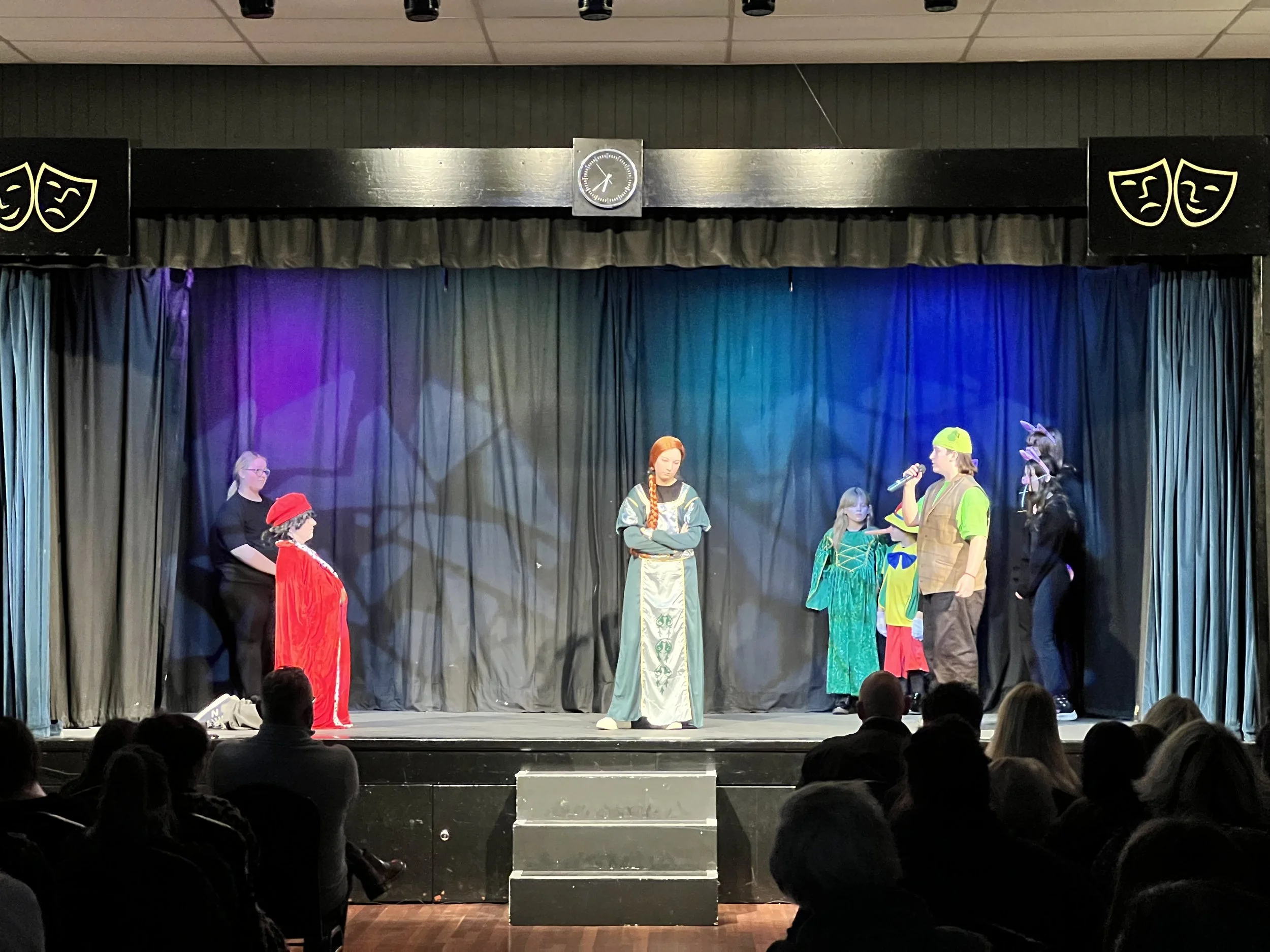 Children on stage performing a play or skit in colorful costumes with an audience watching.