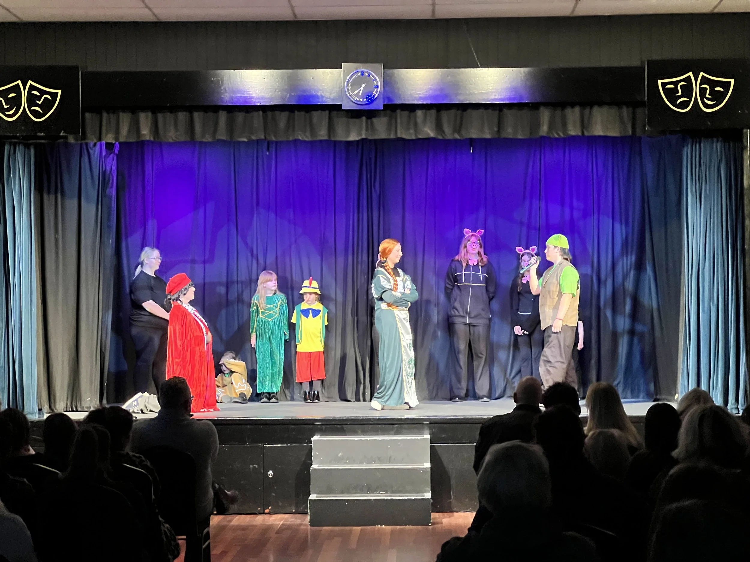Children performing a play on stage with a purple curtain backdrop, wearing colorful costumes including animals and storybook characters, while an audience watches.