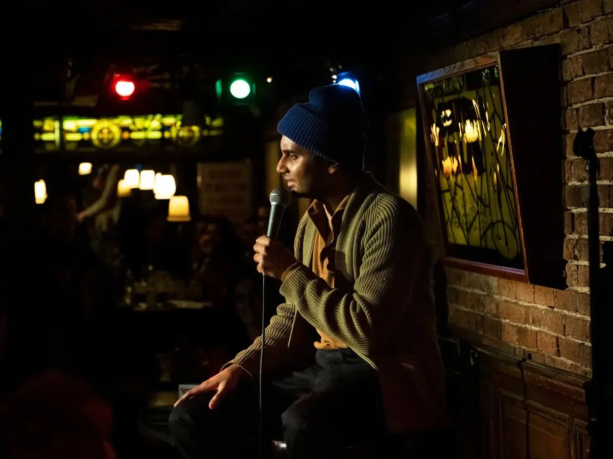 A side view of Aziz Ansari in a night club holding a microphone and looking out into the crowd out of frame.