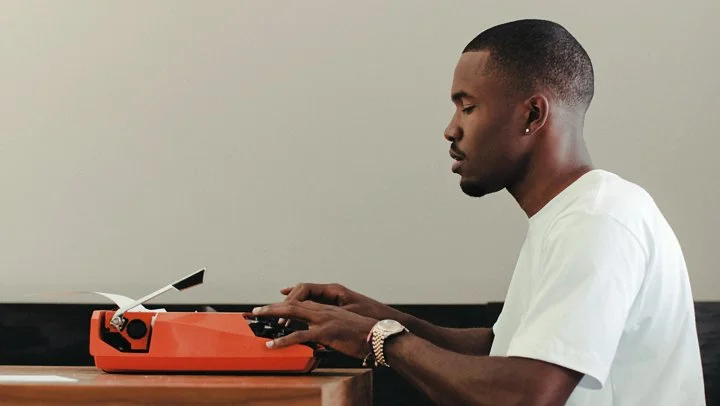 Frank Ocean wearing a white T-shirt, red Polo shorts, and gold wrist watch, sitting a wood table, typing on a Smith Corona Ghia Super G portable orange typewriter.