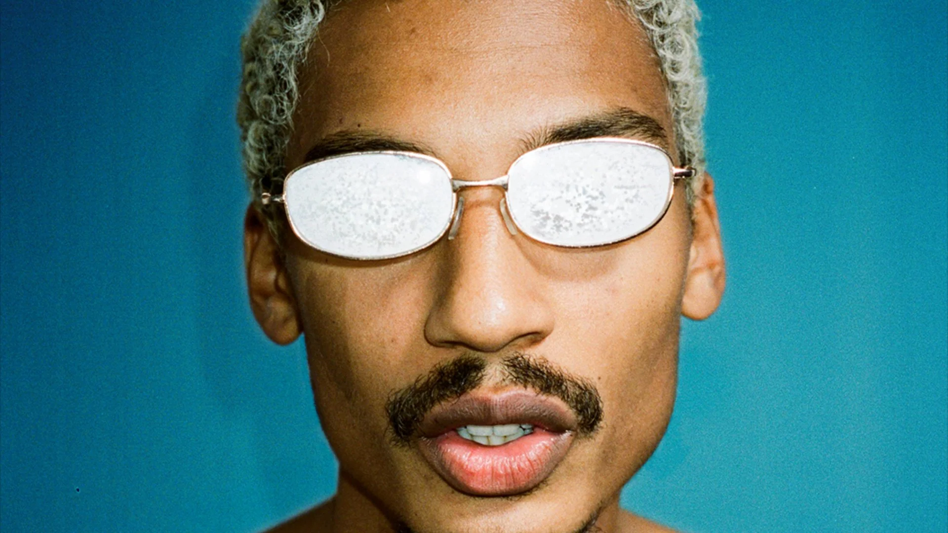 Ysham Jackson with short, blonde hair, whited out glasses, in front of a green-blue background. PRINT ISSUE ONE.