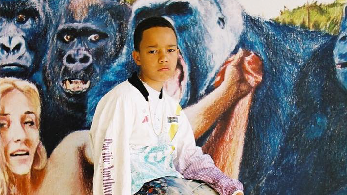 Image of a young boy with short black hair and brown eyes, wearing a gold chain and an Adidas Pacific Northwest long-sleeve T-shirt, smoking a cigarette, and sitting on a stool in front of a mural painting outdoors with a woman and gorillas.
