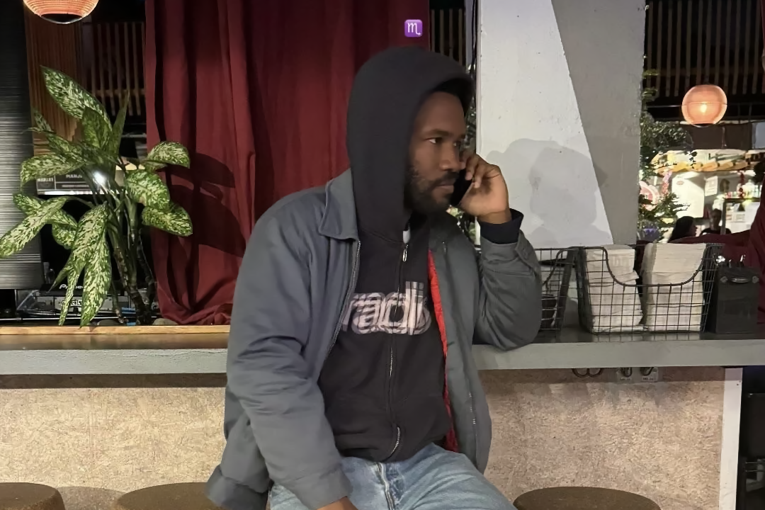 Image of Frank Ocean on the phone, sitting on a bar stool, leaning against the counter. He is wearing a black hoodie with the word 'radio' in white, light blue jeans, and black shoes.