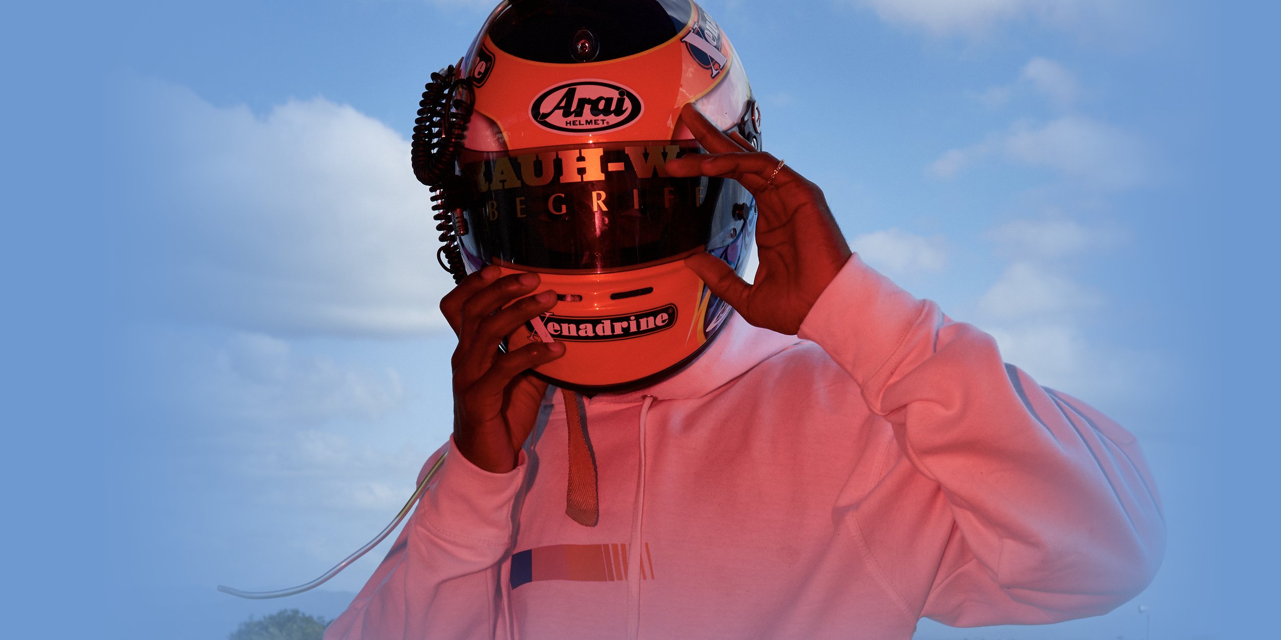 Photo of Frank Ocean wearing a striped hoodie and race helmet branded by RWB and Xenadrine, with his hands at either side and eyes gazing off to the right. Behind him is a racing speedway chicane and a blue sky with clouds.