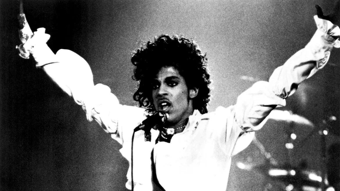 Back and white film photo of Prince singing into a microphone on stage with his arms raised in the air.