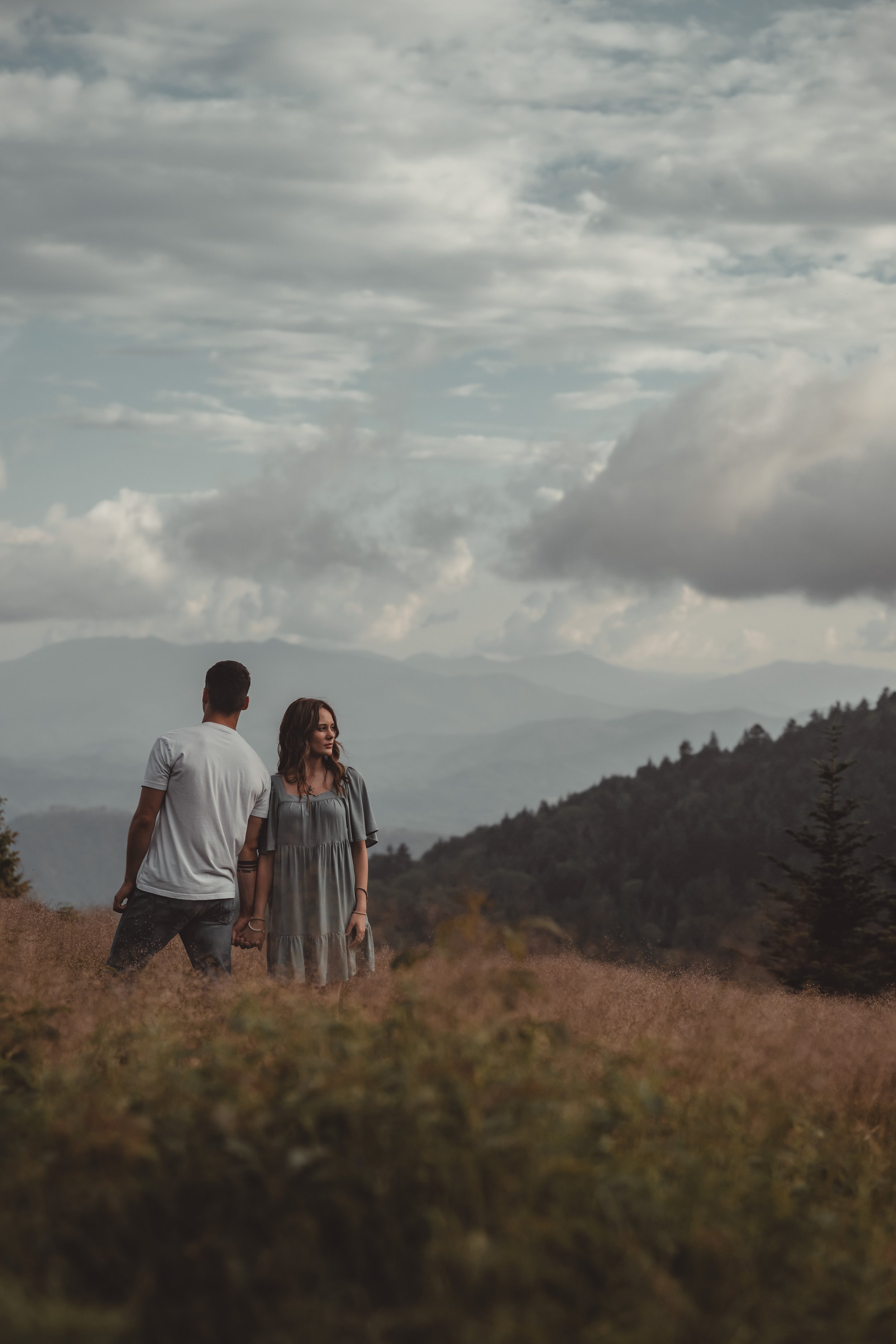 Two people, a man and a woman, standing outdoors on a cloudy day with mountains in the background. The man is wearing a white shirt and has his back to the camera, while the woman is wearing a gray dress and is facing slightly to the right.